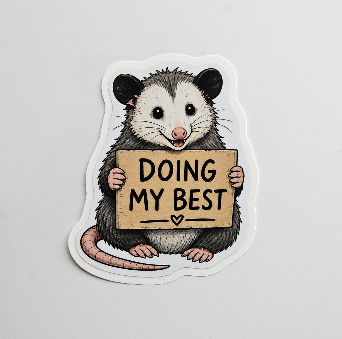 Sticker of an opossum holding a sign that says 'Doing My Best' on a light gray background