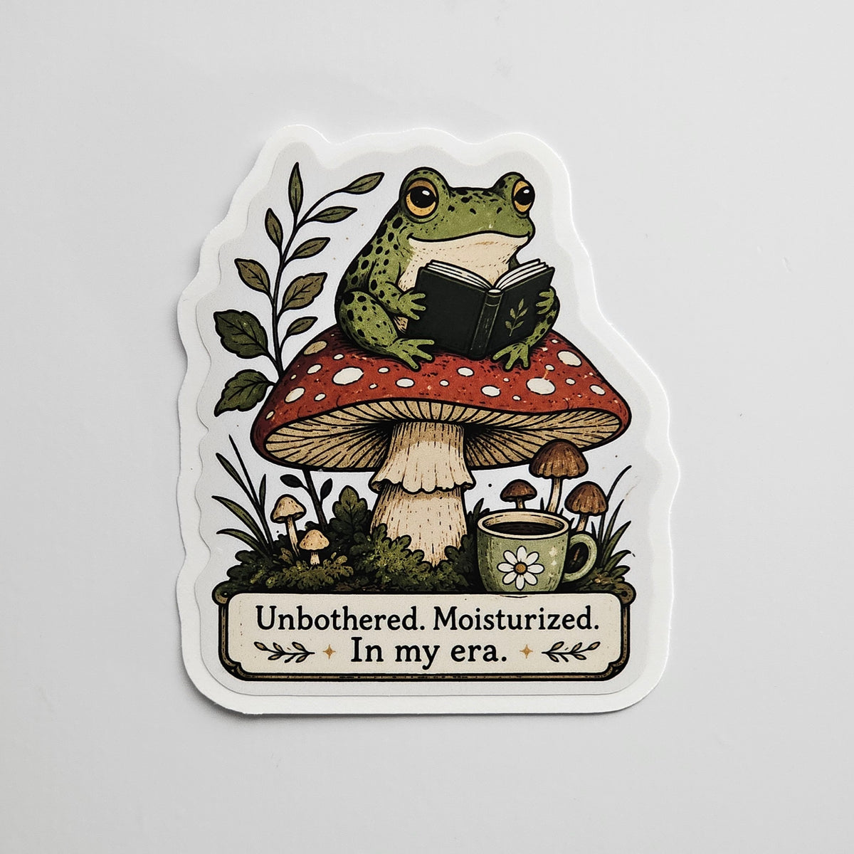 Die-cut sticker with gloss lamination of a cozy frog reading a book while sitting on a mushroom