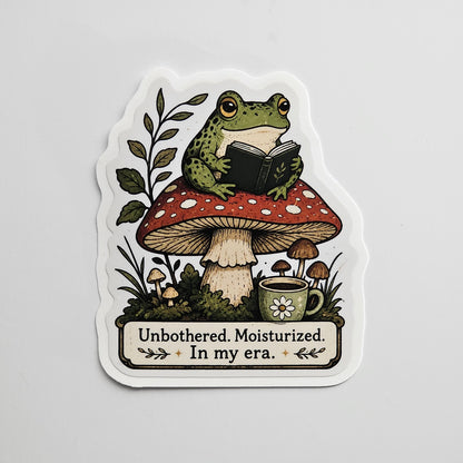 Die-cut sticker with gloss lamination of a cozy frog reading a book while sitting on a mushroom